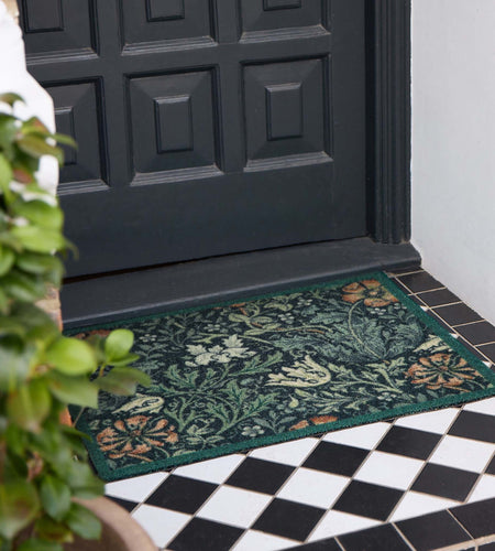 A rectangular floral outdoor doormat in dark green with orange and cream botanical patterns, placed on a tiled entryway in front of a black front door.
