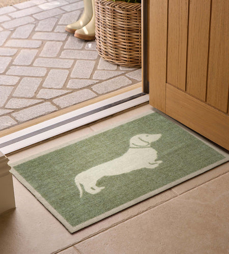 A rectangular green doormat with a white dachshund silhouette, placed in front of an open wooden door on a tiled floor.