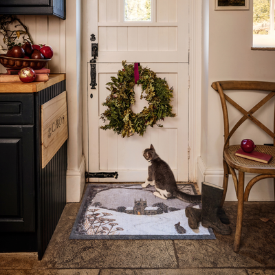 A winter-themed doormat with a snowy landscape and castle design is shown on a stone floor in front of a white door with a green wreath. A gray and white cat sits on the rug, and a wooden chair, dark cabinet, and boots are visible in the cozy entryway setting.