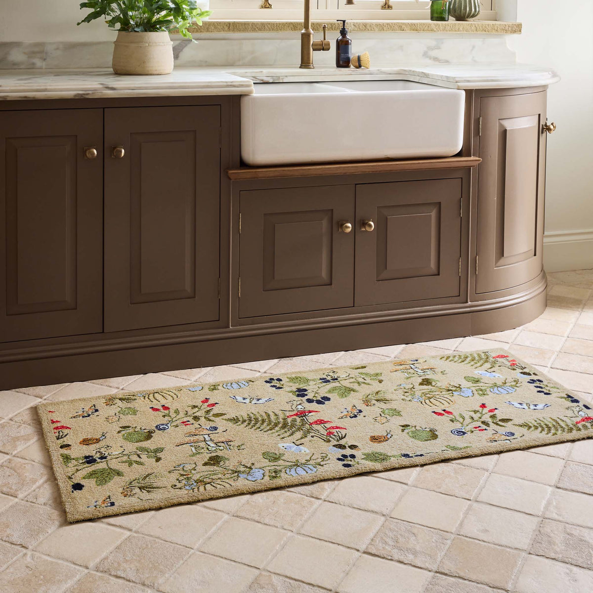 A rectangular beige runner with a colorful embroidered floral and botanical pattern, shown on a tiled kitchen floor in front of a brown vanity and white farmhouse sink.
