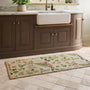 A rectangular beige runner with a colorful embroidered floral and botanical pattern, shown on a tiled kitchen floor in front of a brown vanity and white farmhouse sink.