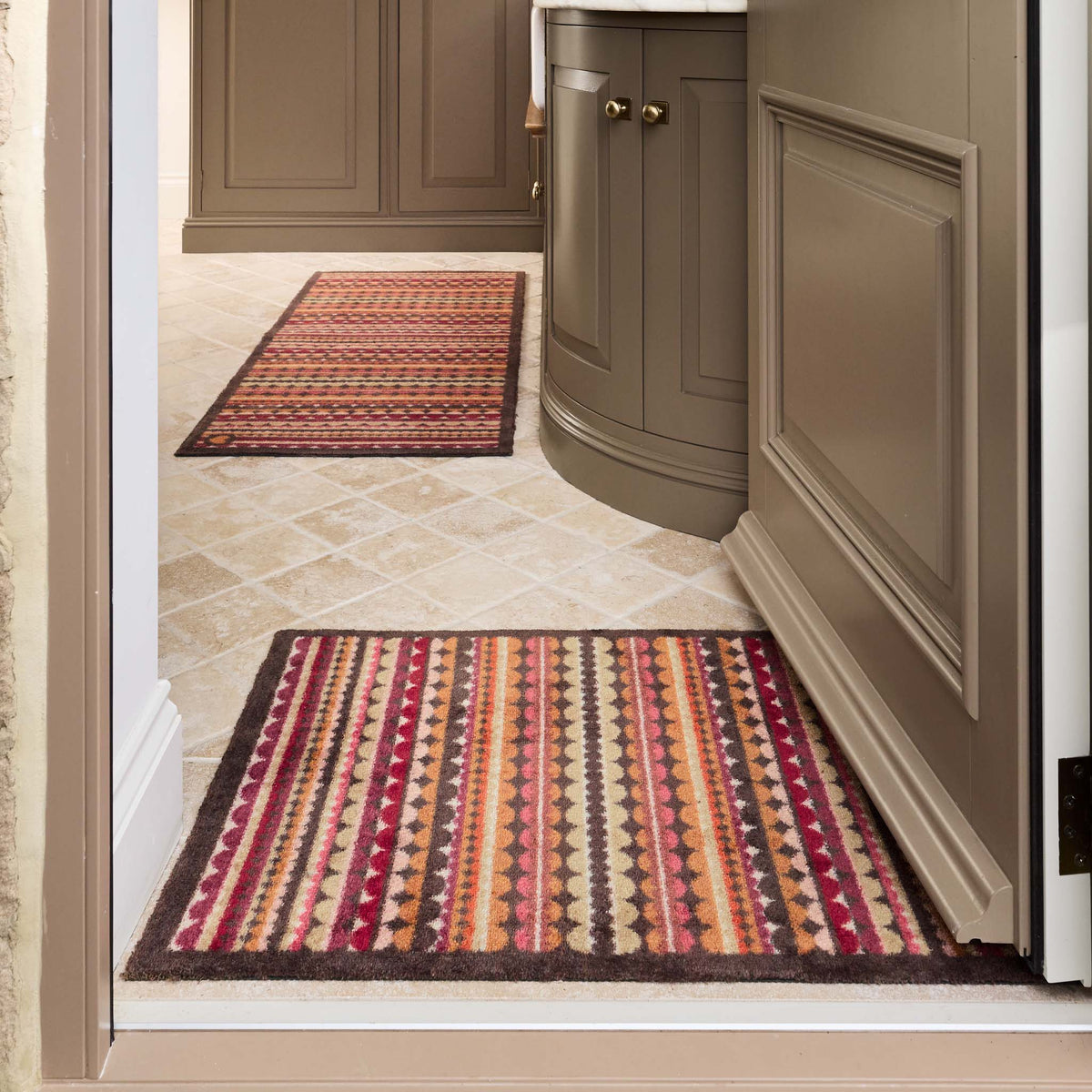 A rectangular doormat with a multicolor Southwestern-style geometric pattern featuring vertical stripes of red, orange, brown, beige, and cream, finished with a dark brown border.