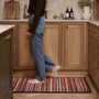 A person walks barefoot across a rectangular woven kitchen mat with a red, beige, and brown striped pattern in a modern kitchen with light oak cabinets.