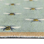 Close-up of a textured area rug featuring a light sage green background with a repeating pattern of yellow and black bumblebees, finished with a beige and white fringed edge.