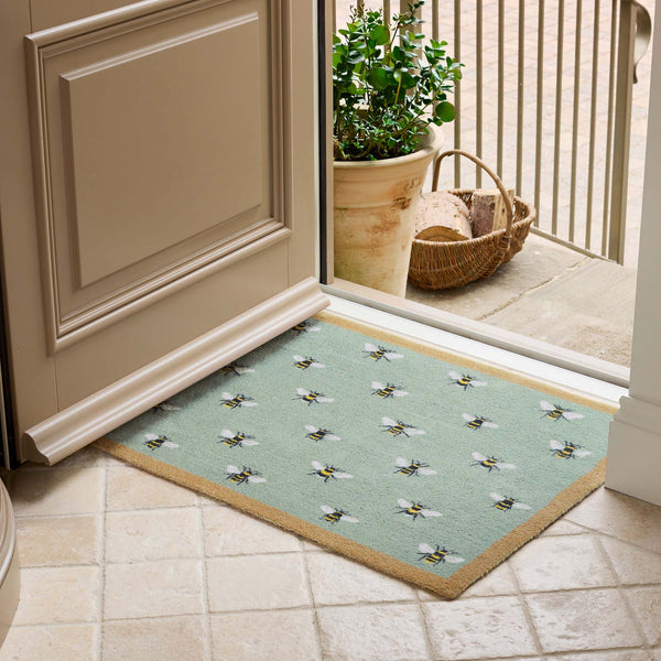 A rectangular entryway doormat with a sage green background, a natural border, and a repeating pattern of small bumblebees, shown on a tiled floor in front of a door.