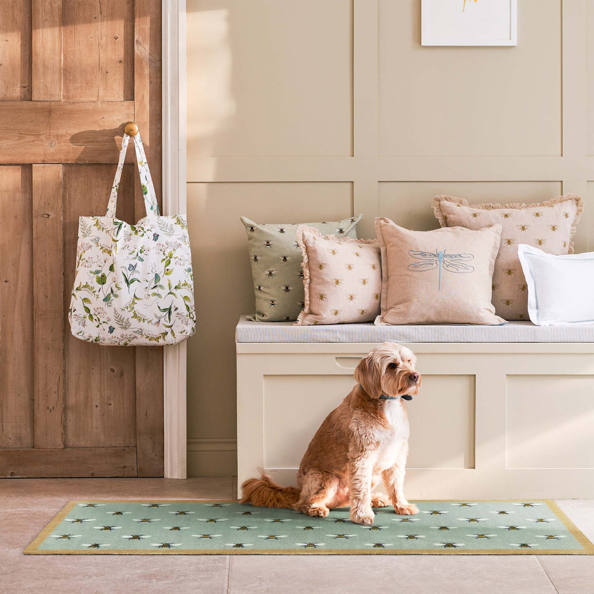 A small tan dog sits on a green patterned mat in a sunlit entryway, with a white tote bag hanging on a wooden door and decorative pillows on a bench behind it.