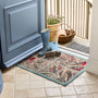 A rectangular doormat with a vintage-style bird panel design in beige, teal, red, and brown tones, placed in front of a blue door. A pair of blue rain boots and a wicker basket sit on the mat at the doorway.