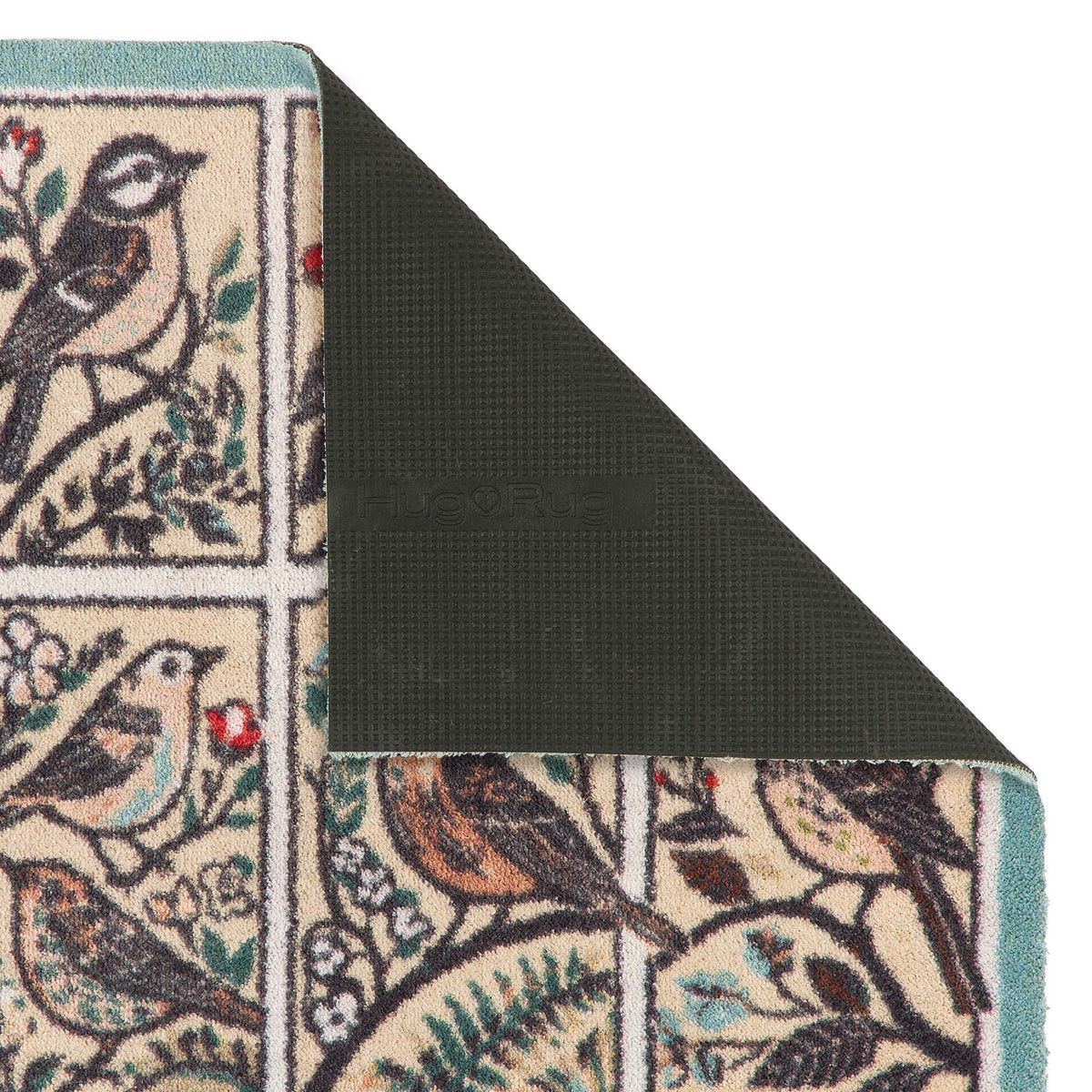 A close-up of a patterned doormat corner showing a bird-and-floral design in cream, teal, brown, and green, with the underside revealing a dark textured backing.
