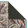 A close-up of a patterned doormat corner showing a bird-and-floral design in cream, teal, brown, and green, with the underside revealing a dark textured backing.
