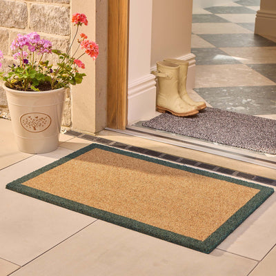 A rectangular coir doormat with a dark green border is placed on a tiled entryway floor in front of a doorway. A potted flower sits on the left, and a pair of cream rain boots rests on a gray mat inside the doorway.