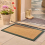 A rectangular coir doormat with a dark green border is placed on a tiled entryway floor in front of a doorway. A potted flower sits on the left, and a pair of cream rain boots rests on a gray mat inside the doorway.