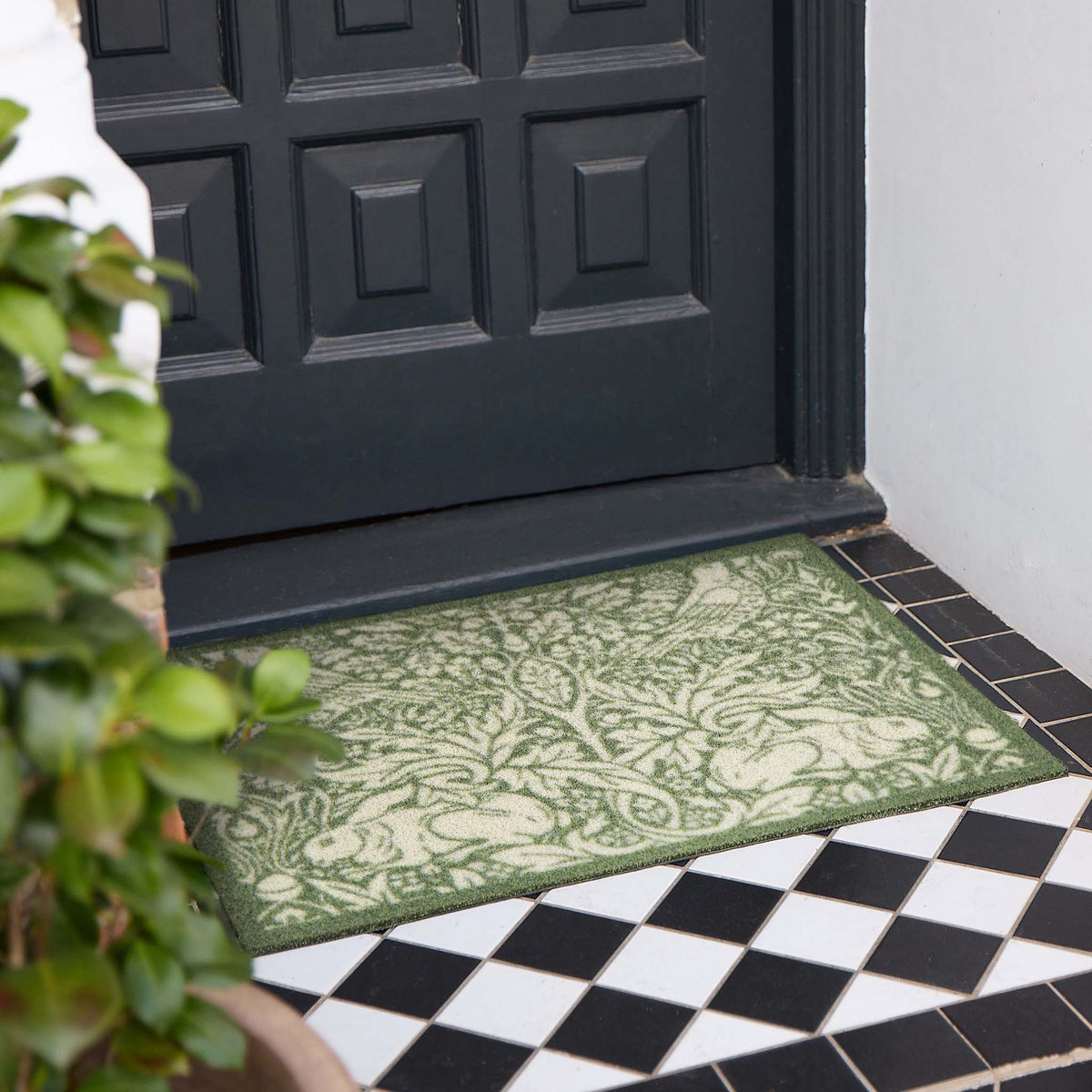 An outdoor rectangular doormat with an ornate green and beige floral pattern, placed in front of a black paneled door on a black and white checkered tile floor.