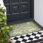 An outdoor rectangular doormat with an ornate green and beige floral pattern, placed in front of a black paneled door on a black and white checkered tile floor.