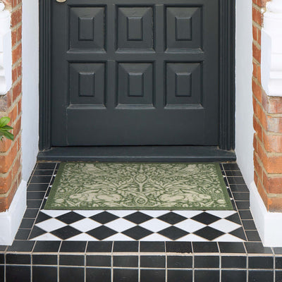 A rectangular outdoor doormat with an ornate green and beige damask pattern, placed on a tiled porch step in front of a black front door.