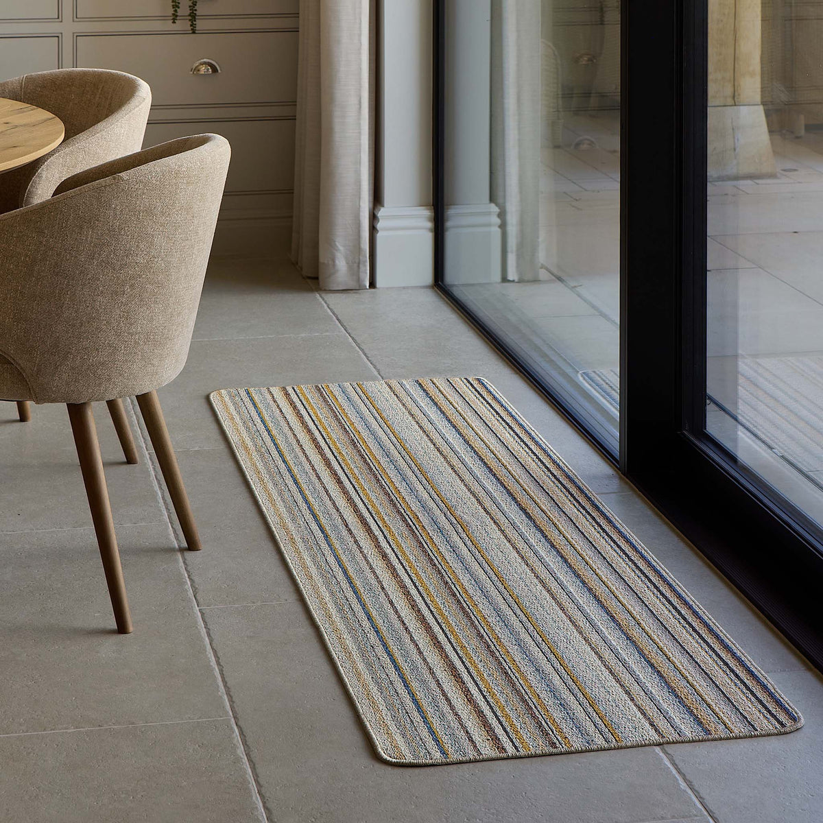 A rectangular woven runner rug with vertical stripes in beige, blue, and yellow, placed on a light gray tiled floor next to a glass patio door in a modern dining room setting.