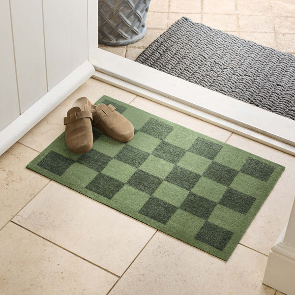 A green checkered doormat placed at a doorway on a light tile floor, with a pair of tan clogs on the front and a gray patterned mat visible beyond the threshold.