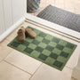 A green checkered doormat placed at a doorway on a light tile floor, with a pair of tan clogs on the front and a gray patterned mat visible beyond the threshold.