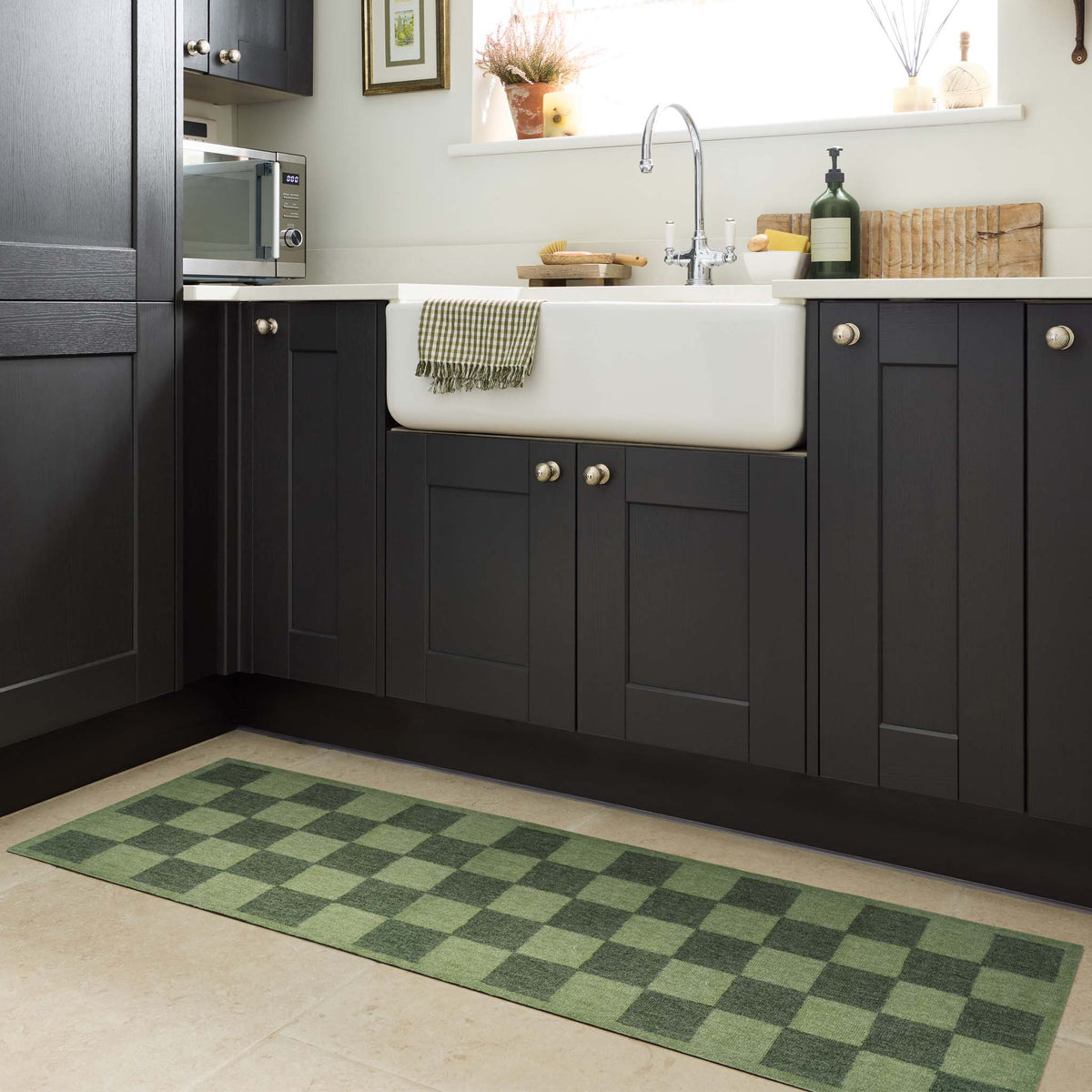 A green and dark green checkered kitchen runner on a light beige tiled floor in a modern kitchen setting with dark cabinets and a white farmhouse sink.