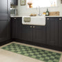 A green and dark green checkered kitchen runner on a light beige tiled floor in a modern kitchen setting with dark cabinets and a white farmhouse sink.