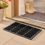 A black rubber entryway mat with integrated drainage channels, shown on a tiled floor at a doorway. A pair of cream rain boots and a gray doormat are visible on the porch step behind it.