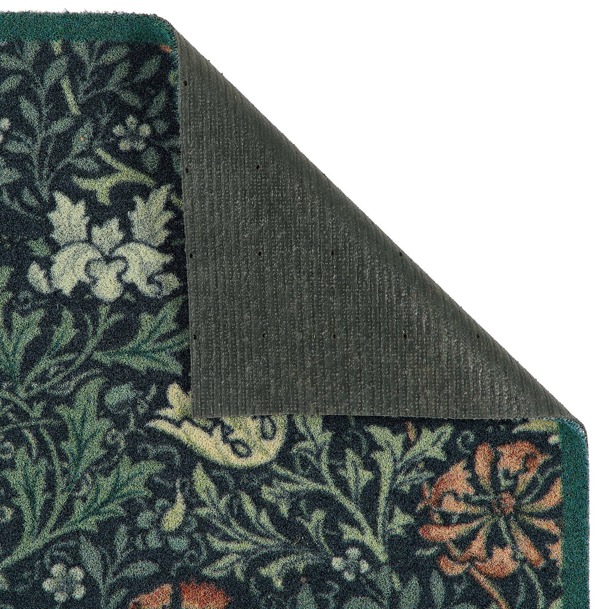 A close-up of a patterned doormat corner with the top-left corner folded back to show the dark gray backing. The rug features a dark navy blue background with a detailed botanical and floral pattern in green, cream, and rust orange.