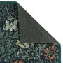 A close-up of a patterned doormat corner with the top-left corner folded back to show the dark gray backing. The rug features a dark navy blue background with a detailed botanical and floral pattern in green, cream, and rust orange.