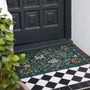 A rectangular outdoor doormat with an intricate floral and botanical pattern in dark green, beige, and rust orange, placed on a black-and-white checkered tiled floor in front of a dark paneled door.