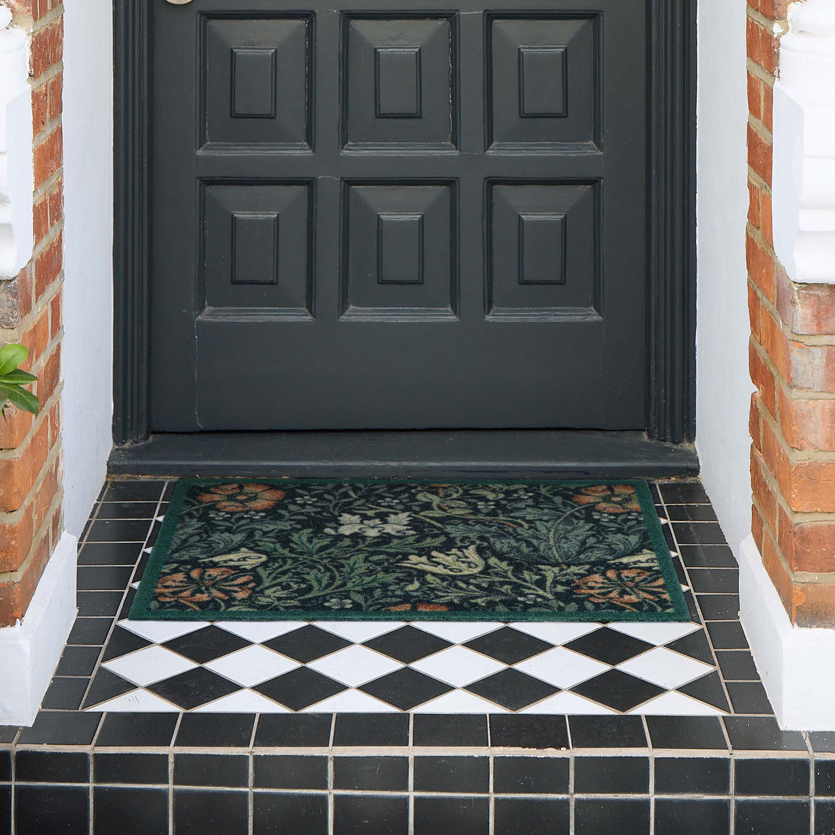 A rectangular floral outdoor doormat in dark green with orange and cream botanical patterns, placed on a tiled entryway in front of a black front door.