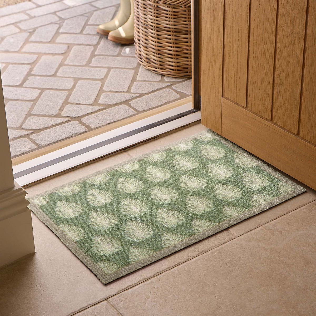 A rectangular entryway doormat with a green and beige palm leaf pattern, positioned on a tiled floor in front of an open wooden door.