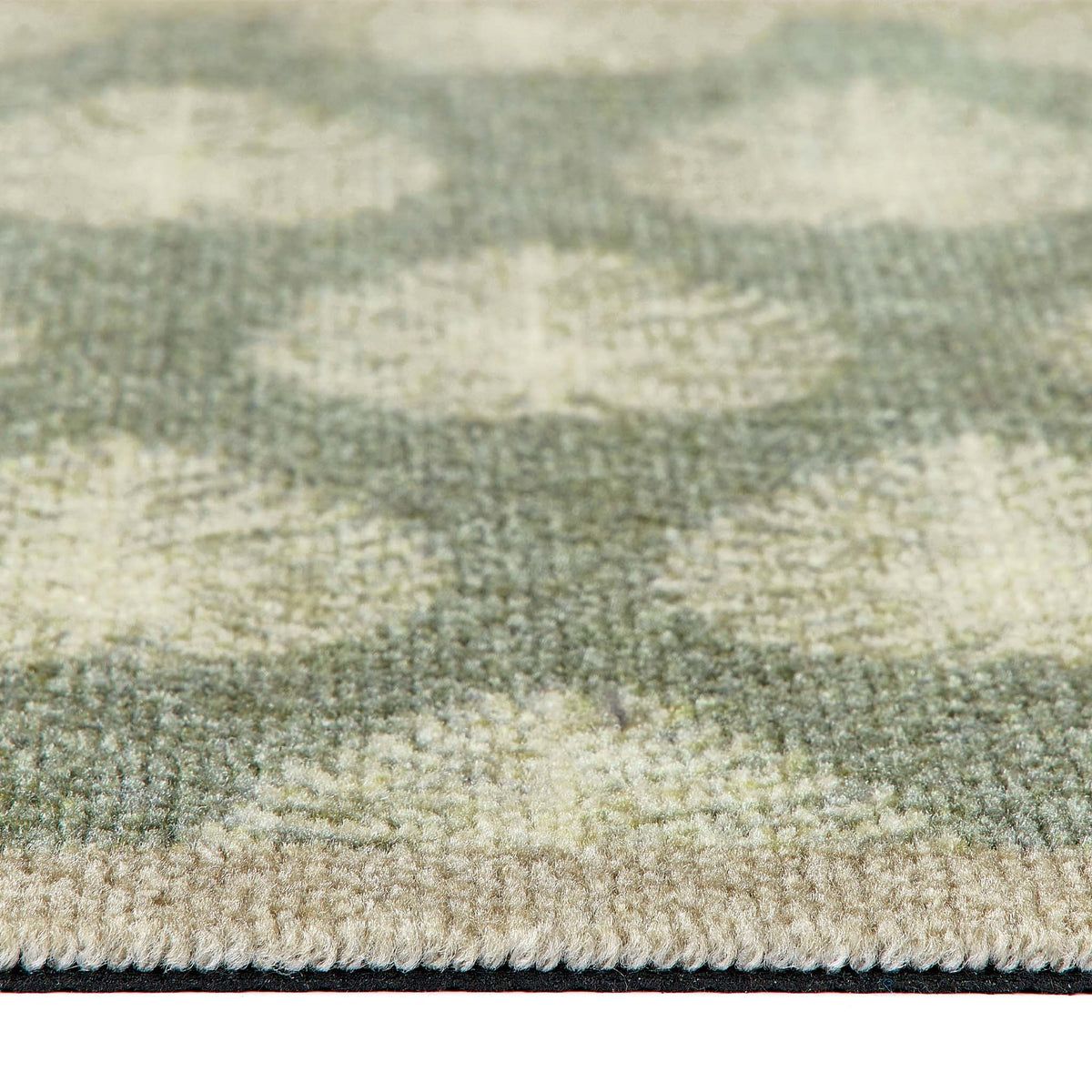 Close-up of a textured doormat with a leaf pattern in sage green, beige, and cream, showing the pile texture and fringed edge.