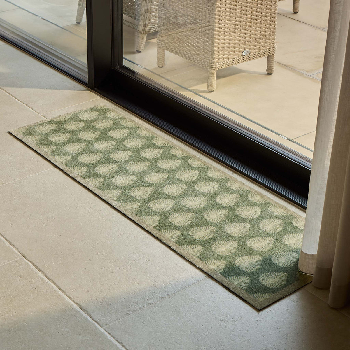 A rectangular runner with a green and beige geometric pattern, positioned on a light stone patio floor in front of a glass sliding door.