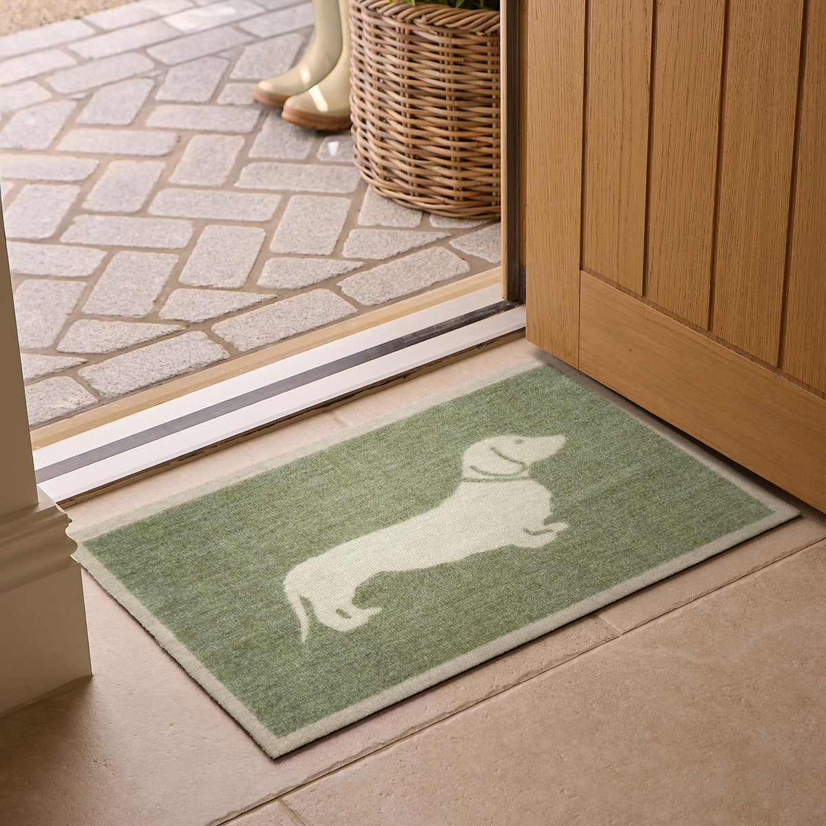 A rectangular green doormat with a white dachshund silhouette, placed in front of an open wooden door on a tiled floor.