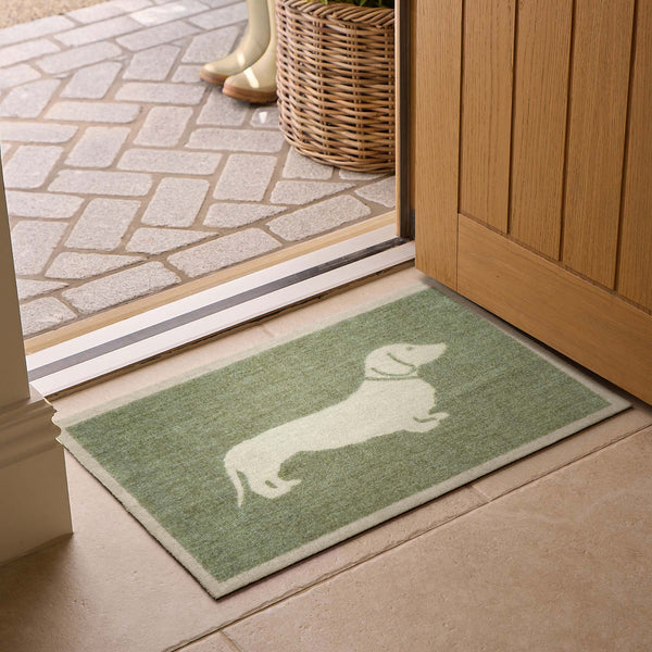 A rectangular green doormat with a white dachshund silhouette, placed in front of an open wooden door on a tiled floor.