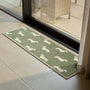 A rectangular indoor runner with a sage green background and a repeating pattern of white dachshund dog silhouettes, placed on a light stone tile floor in front of a glass patio door.