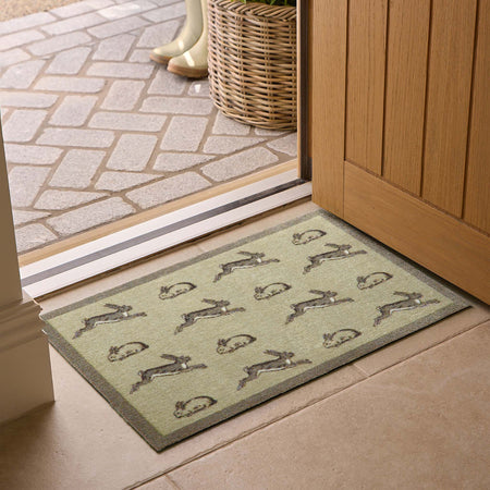 A rectangular entryway doormat with a repeating rabbit pattern, placed in front of a sliding glass door on a tiled floor. The mat has a muted sage green base with gray and brown rabbits and a darker gray border.