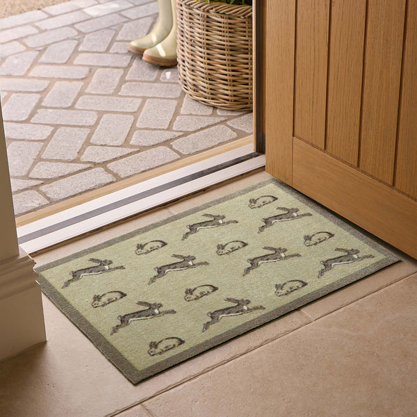 A rectangular entryway doormat with a repeating rabbit pattern, placed in front of a sliding glass door on a tiled floor. The mat has a muted sage green base with gray and brown rabbits and a darker gray border.
