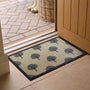 A rectangular entryway doormat with a repeating tree pattern, shown on the floor at an open doorway. The mat has a dark grey border and a light beige background with navy tree motifs.