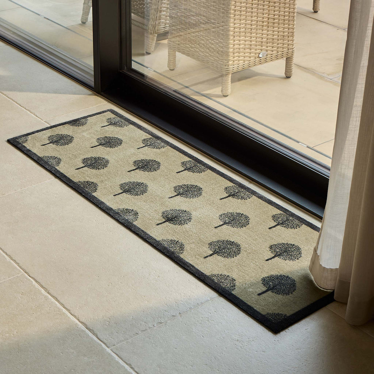 A rectangular indoor runner with a black tree pattern on a beige background, placed on a light stone tile floor in front of a glass sliding door.