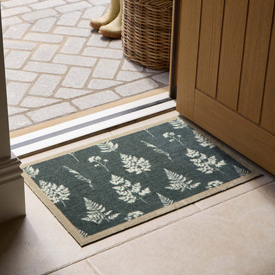 A rectangular entryway doormat with a dark green background and a white fern pattern, placed on the floor in front of an open wooden door. A wicker basket and boots are visible outside.