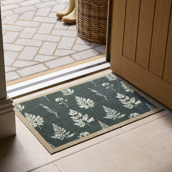 A rectangular entryway doormat with a dark green background and a white fern pattern, placed on the floor in front of an open wooden door. A wicker basket and boots are visible outside.