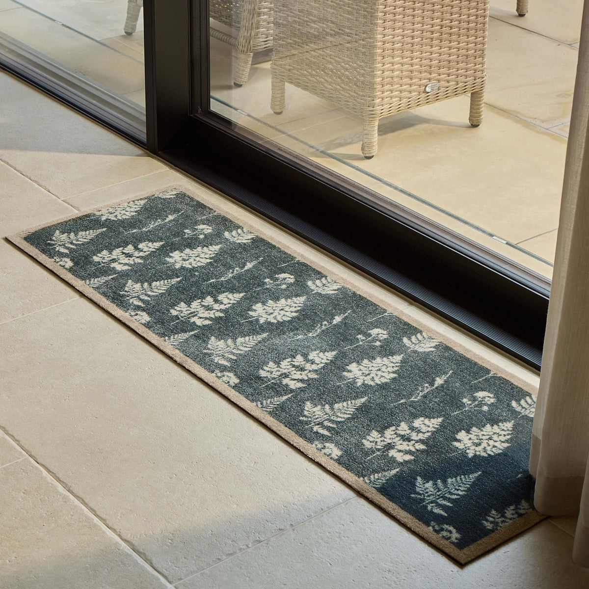 A rectangular runner with a dark green background and a repeating white leaf pattern, placed on a light stone tile floor in front of a glass sliding door.