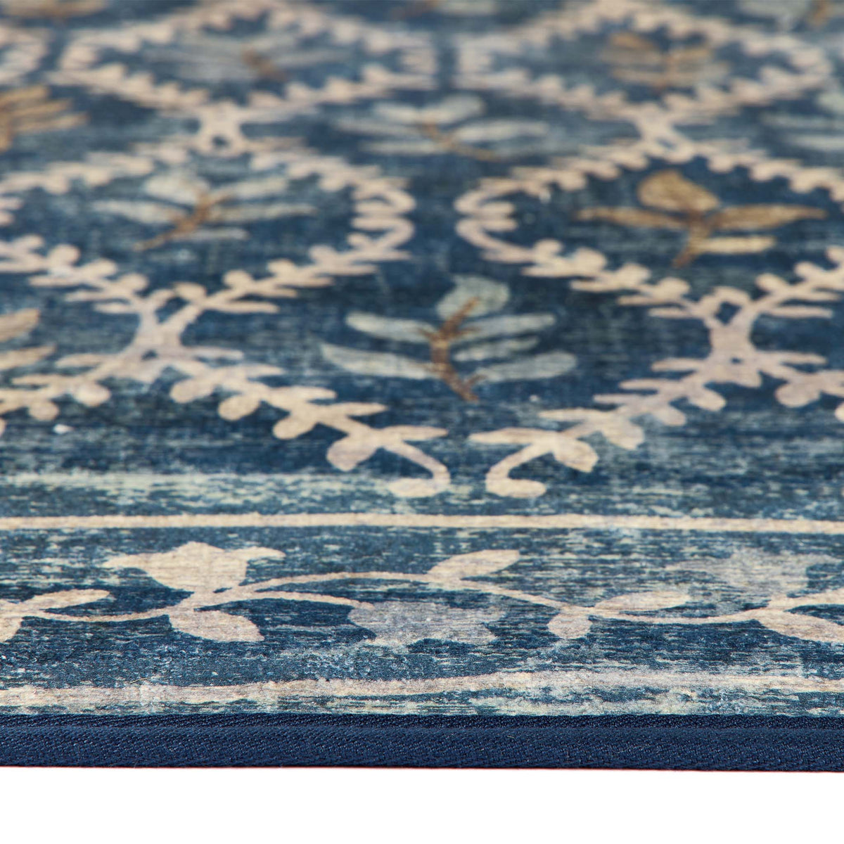A close-up of a blue area rug with a distressed floral and leaf pattern in cream and muted brown tones.