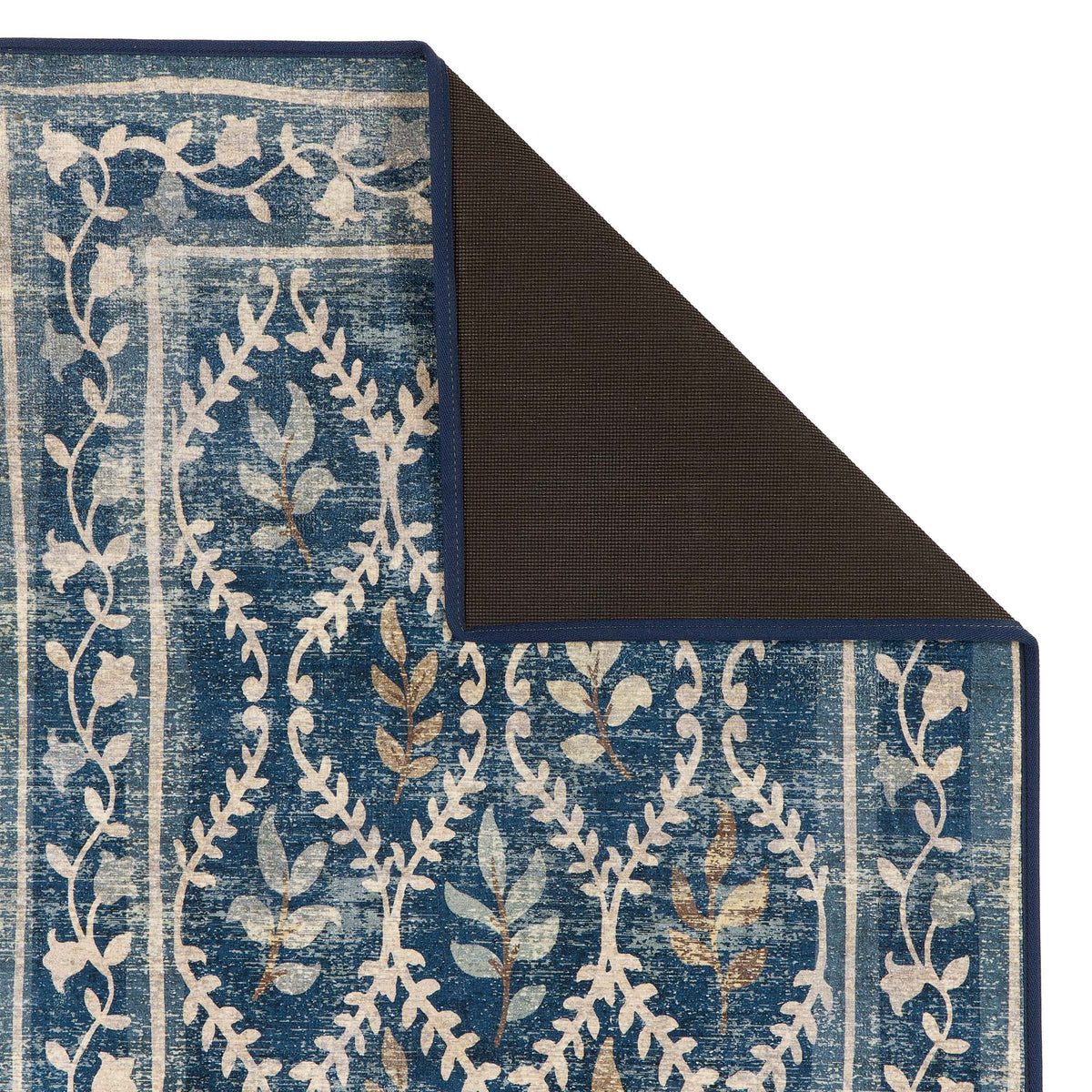 A close-up of a patterned area rug with a folded corner showing the dark backing, featuring a blue and cream floral vine design with subtle gold accents.