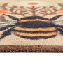 Close-up of a doormat featuring a stylized bumblebee design in black, mustard yellow, and orange-brown on a warm cream background.