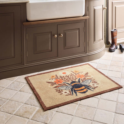 A rectangular beige doormat with a brown border and a central bumblebee design surrounded by orange flowers, placed on a tiled kitchen floor in front of a dark wood vanity.