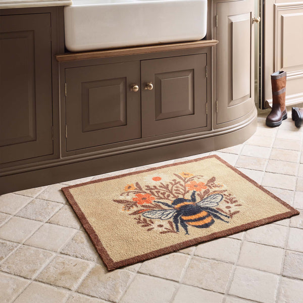 A rectangular beige doormat with a brown border and a central bumblebee design surrounded by orange flowers, placed on a tiled kitchen floor in front of a dark wood vanity.