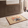 A rectangular beige doormat with a brown border and a central bumblebee design surrounded by orange flowers, placed on a tiled kitchen floor in front of a dark wood vanity.