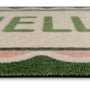Close-up of a textured doormat with a cream background, green lettering, and a green and pink patterned border.