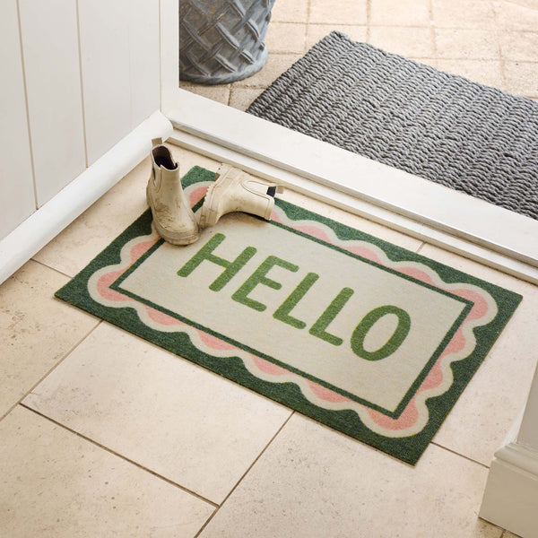 A rectangular entryway doormat with the word “HELLO” in green, bordered in dark green and pink scalloped trim, placed on a tiled floor at a doorway with a pair of off-white boots resting on it.
