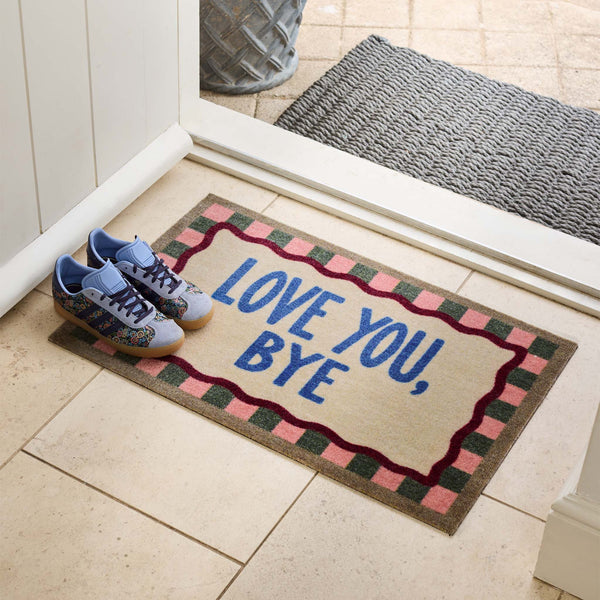A rectangular doormat with the words “LOVE YOU, BYE” in blue sits on a tiled entryway floor next to a pair of blue sneakers. The mat has a beige center, a pink and green checkered border, and a dark red wavy inner border.
